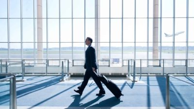 A man in a dark blue suit walks through an airport past floor-to-ceiling windows with a Qantas plane flying in the distance