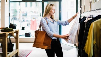 A young woman in a shop wearing black pants and a blue blouse looks at a white shirt off the rack