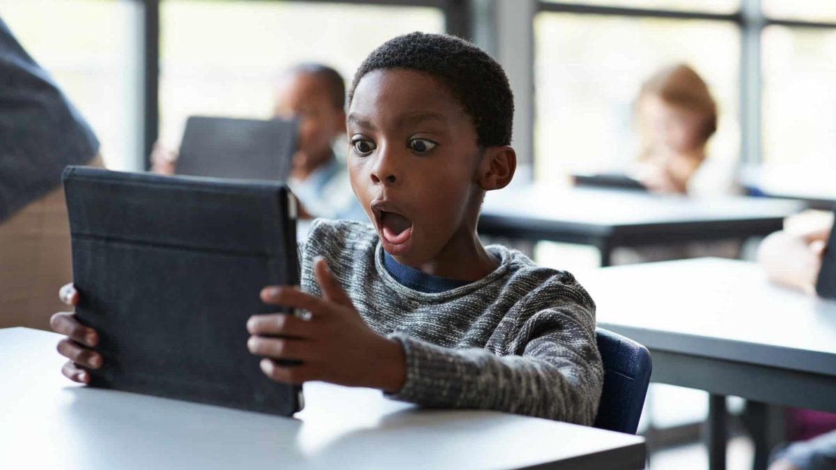 A young schoolboy sits at his desk in a classroom with awe in his face as he looks at his ipad