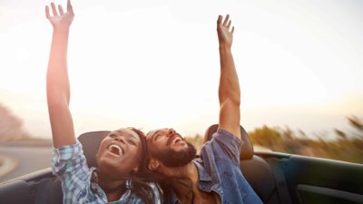 A young couple in the back of a convertible car each raise a single arm in the air whilst enjoying a drive along the road.