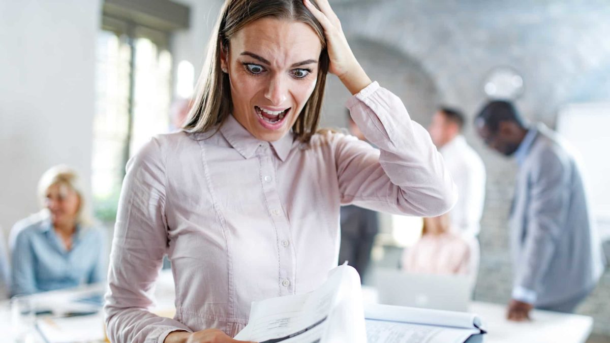 A young businesswoman looks shocked at what she's reading on the paperwork in her hand, with colleagues in the office in the background.
