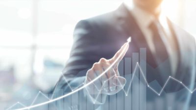 a business person in a suit and tie directs a pointed finger upwards with a graphic of a rising bar graph and an arrow heading upwards in line with the person's finger.