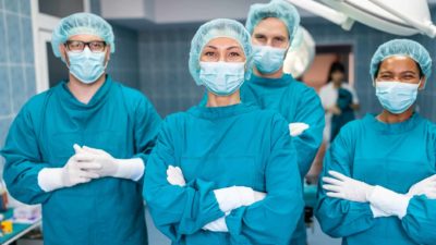 a group of surgeons in full surgery dress including masks, gloves and head coverings stands together with arms folded and smiling eyes as if happy with the outcome of their efforts.