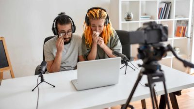 A couple sit at a desk with tissues and tears in their eyes while they look at a laptop computer screen with a camera set up in the foreground suggesting they are making a video.