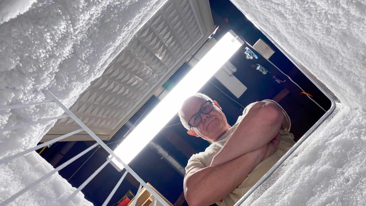 a man stands with arms folded and an unhappy expression on his face looking up from within an iced-up chest freezer in need of defrosting. It's as though the picture has been taken from within the freezer.