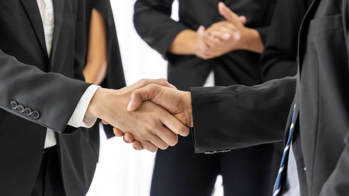 two businessmen shake hands in a close up mid-level shot with other businesspeople looking on approvingly in the background.