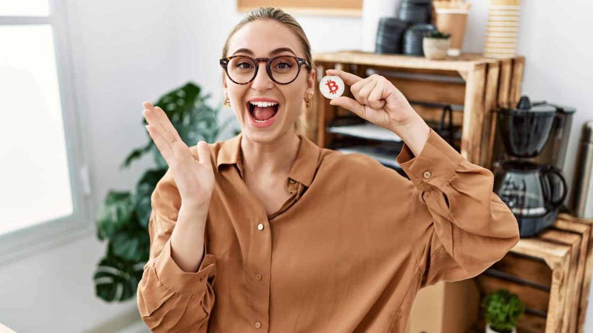 A young woman wearing work wear in an office setting has a lively, happy, open-mouthed expression of joy while holding a bitcoin token.