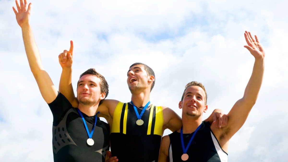 three men stand on a winner's podium with medals around their necks with their hands raised in triumph.
