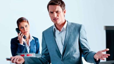 an angry man in a suit stands with his hands outstretched in a questioning gesture of annoyance and displeasure while an airport check in attendant is on the telephone in the background.