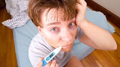 a man with a pale face and bags under his eyes sits on the end of his bed with a thermometer in his mouth and a hand to his dishevelled head.