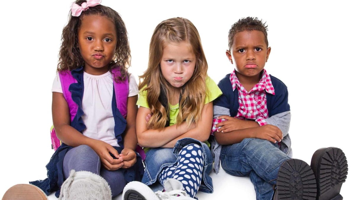 three children in fashionable clothes sit in a row together with sad looks on their faces as though they hae been told not to do something or been curtailed from playing.