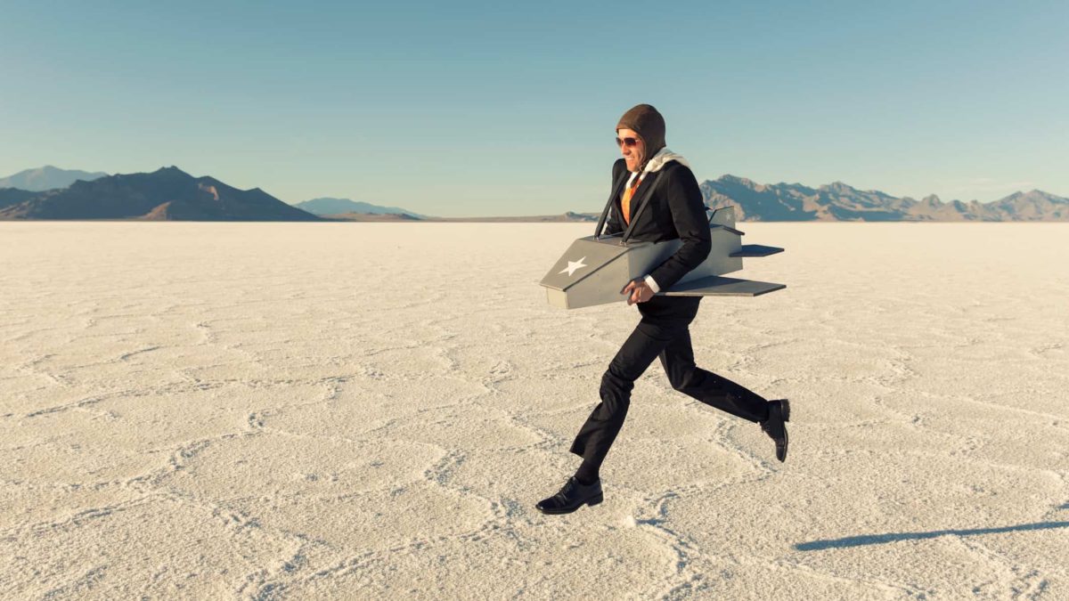 a man wearing an old-fashioned aviation leather head covering and goggles and with a cardboard plane shape around his waist runs along the ground against a barren, desert background.