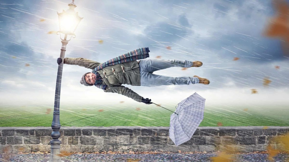 a man blown off his feet sideways hangs on with one hand to a lamp post with an inside out umbrella in his other hand as he is lashed by wind and rain with a grey cloudy sky background.