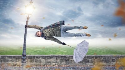 a man blown off his feet sideways hangs on with one hand to a lamp post with an inside out umbrella in his other hand as he is lashed by wind and rain with a grey cloudy sky background.