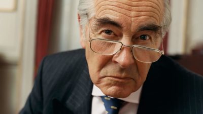 a man in a business suit has a stern look on his face as he leans forward and peers over his glasses.