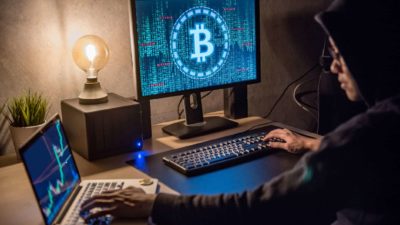 A young male Bitcoin investor sits as his desk in front of a monitor with the bitcoin logo on it and a laptop next to him showing a graph of cryptocurrency stocks