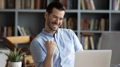 A man in a blue collared shirt sits at his desk doing a single fist pump as he watches the Appen share price rise on his laptop