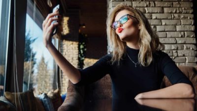 A smug executive woman wearing glasses and red lipstick blows a kiss to herself as she takes a selfie.