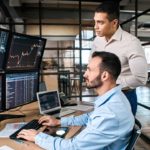 Two male ASX 200 analysts stand in an office looking at various computer screens showing share prices