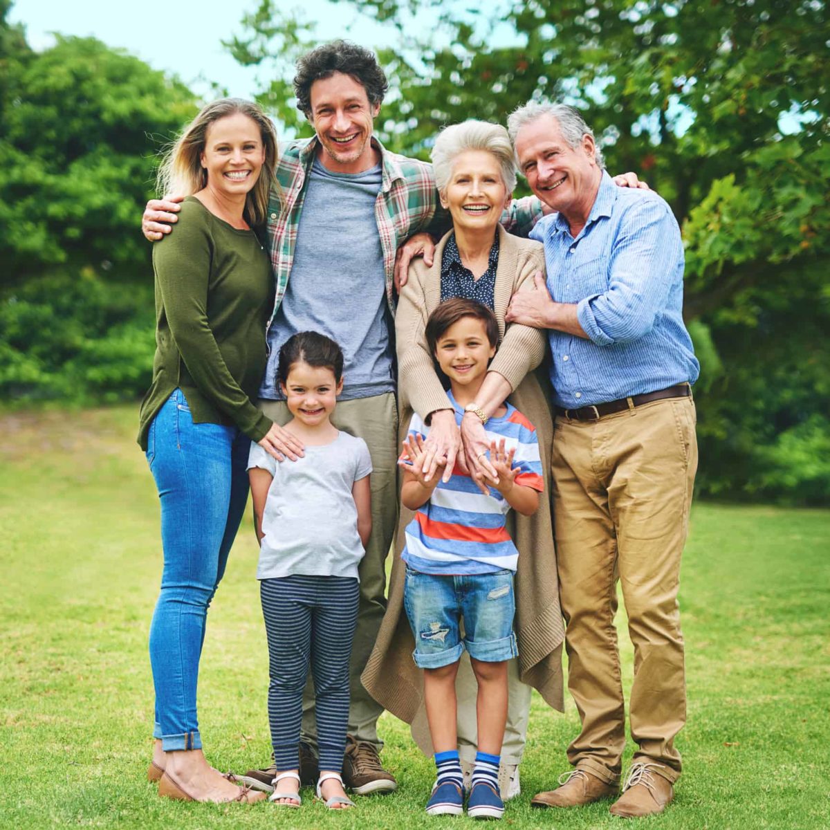 Three generations of a family, grandparents, parents and two children, pose lovingly together on grass with trees in the background.