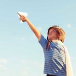 A kid wearing a pilot helmet holds a paper plane up to the sky.