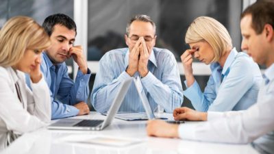 a group of business people sit dejectedly around a table, each expressing desolation, sadness and disappointment by holding their head in their hands, casting their gazes down and looking very glum.