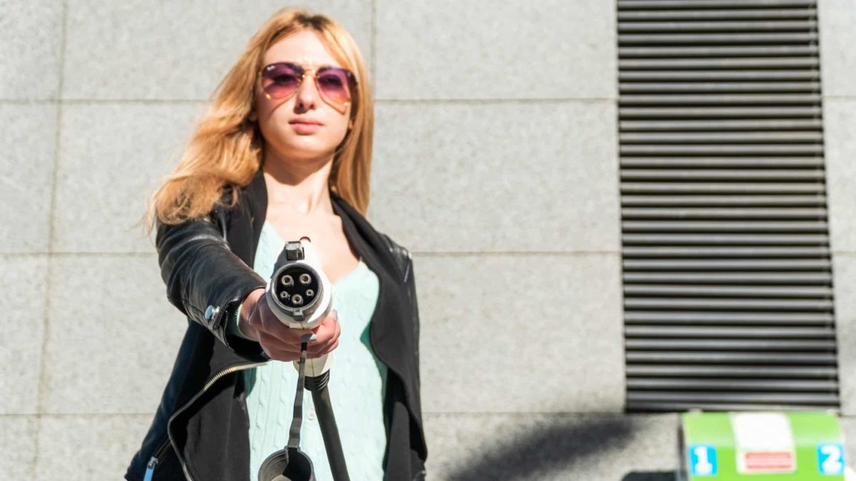 a woman holds out an electric vehicle charger with a satisfied look on her face behind cool sunglasses.