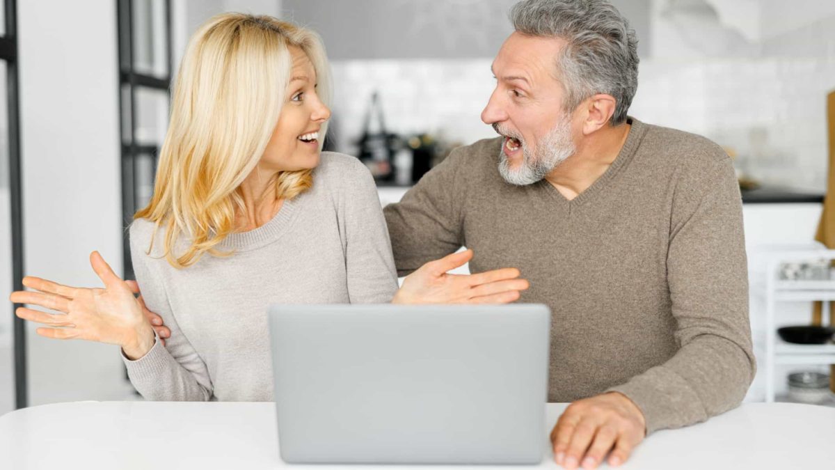 A man and woman sit next to each other looking at each other and feeling excited and surprised after reading good news about their shares on a laptop.