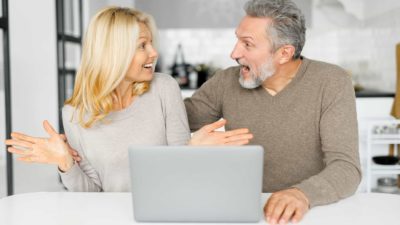 A man and woman sit next to each other looking at each other and feeling excited and surprised after reading good news about their shares on a laptop.