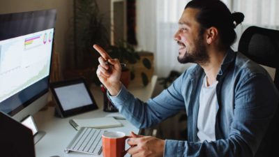 An investor looks happy holding a finger to his computer screen while holding a coffee cup in a home office scenario.