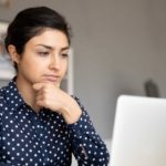 A young woman sits at her desk in deep contemplation with her hand to her chin while seriously considering information she is reading on her laptop