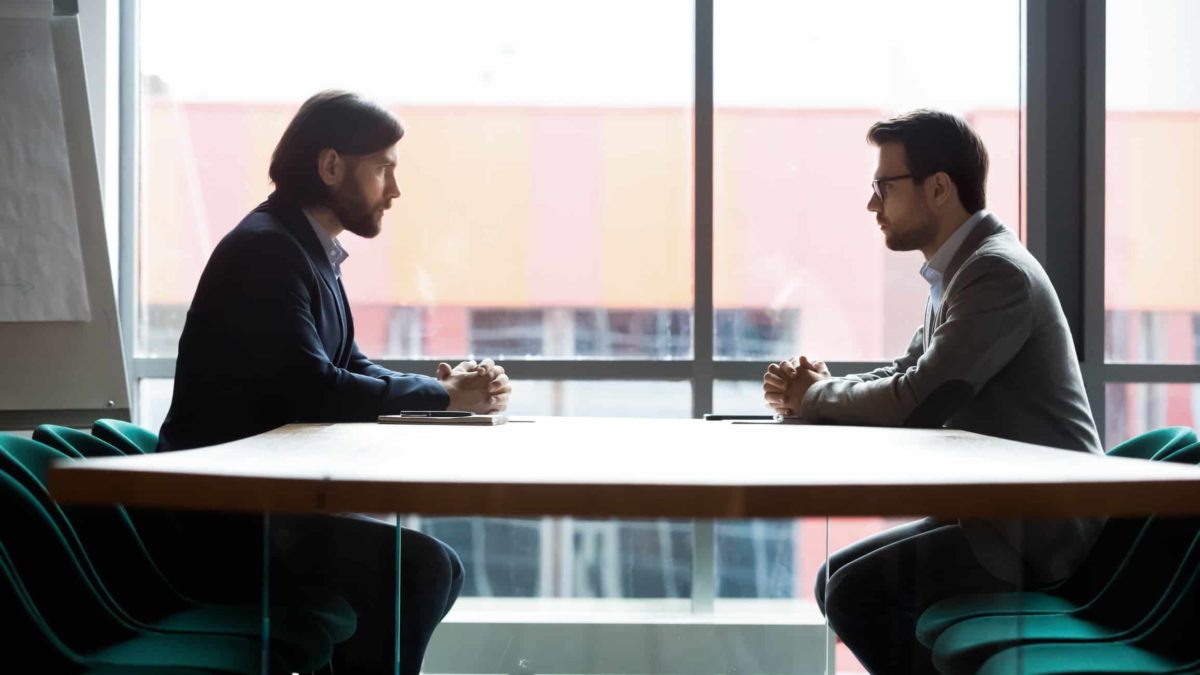 two business men sit across from each other at a negotiating table. with a large window in the background.