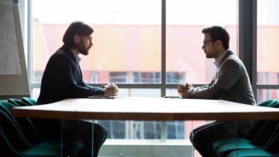 two business men sit across from each other at a negotiating table. with a large window in the background.