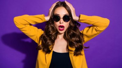 an attractive model-like woman holds her hands to her head and gives a shocked an exasperated wide-mouthed expression as though she is hearing unexpected news.