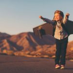 a young girl wearing a set of airplane wings stands on a tarmac with hands in the air and an excited look on her face as though she is about to take off.