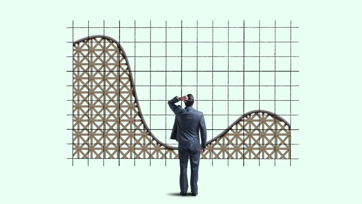A man in a business suit scratches his head looking at a graph that started high then dips, then starts to go up again like a rollercoaster.