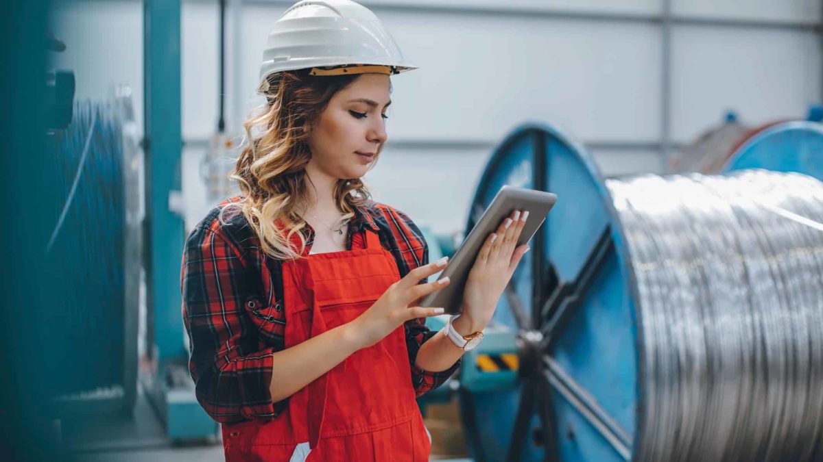 a woman wearing a hard hat and high visibility vest checks her device in front of a large spool of steel cable.