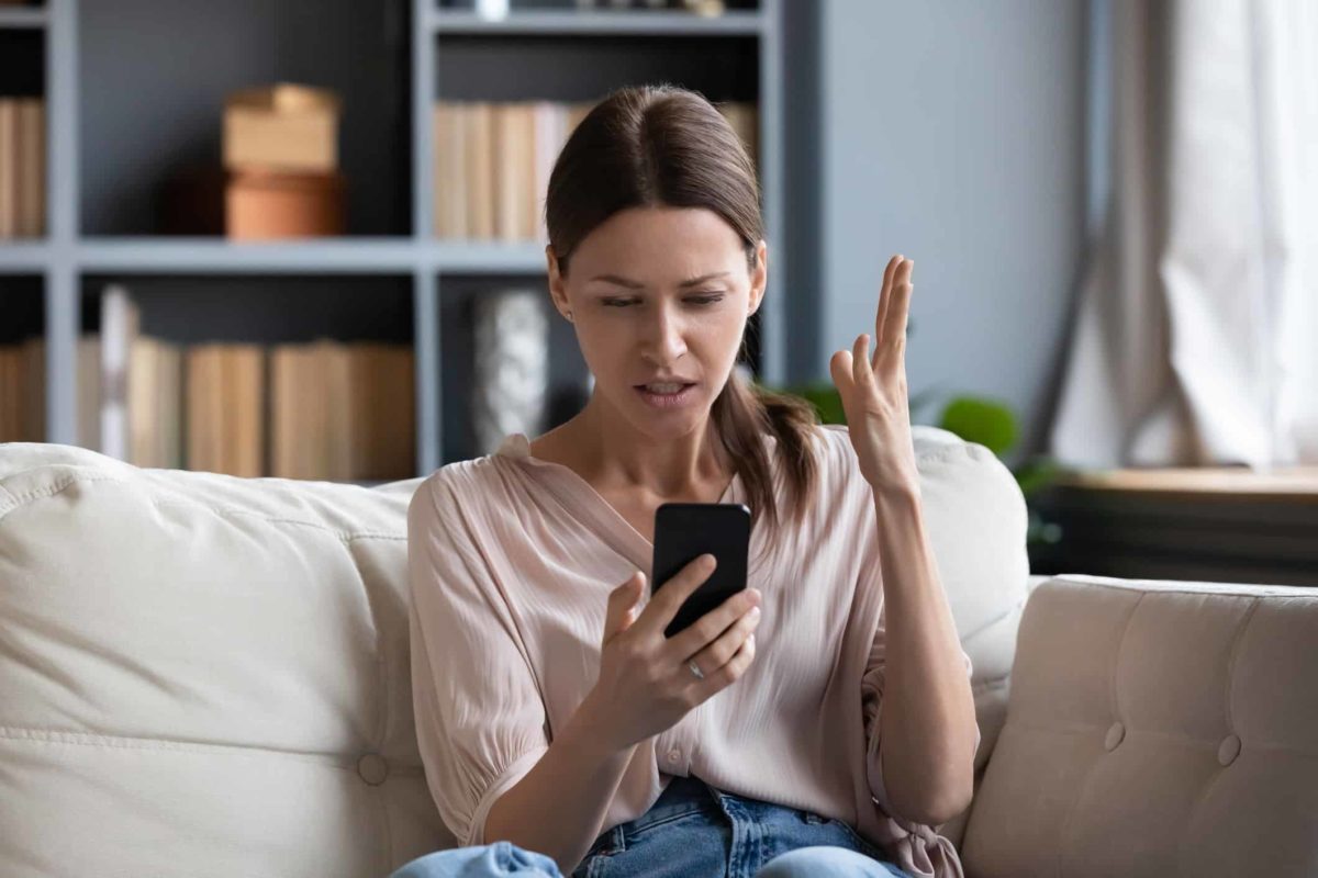A woman sits on her lounge looking stressed and surprised while reading news on her phone.