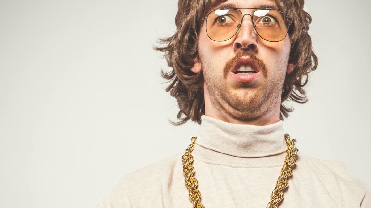 A man wearing 70s clothing and a big gold chain around his neck looks a little bit unsure.