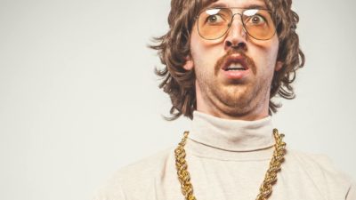 A man wearing 70s clothing and a big gold chain around his neck looks a little bit unsure.