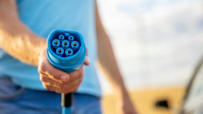 a headless person holds an electric vehicle charger front on to the campera so its plug points are visible, forming the central focal point for the shot.