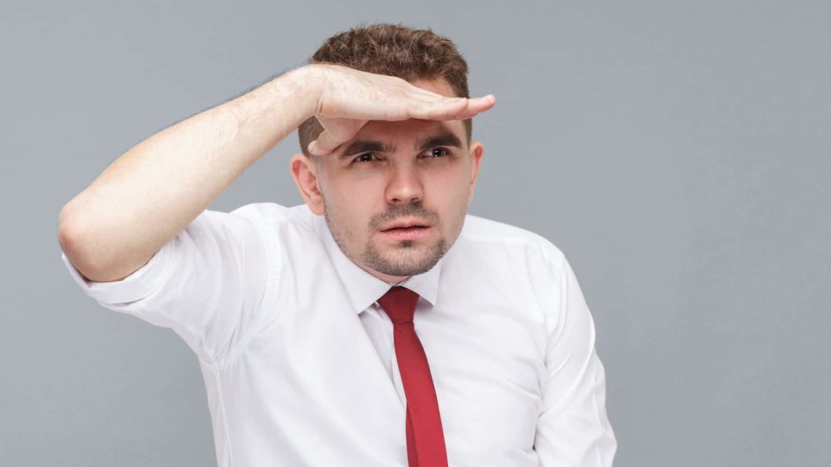 a man in a shirt and tie looks to the horizon holding his hand above his eyes as if to shield the sun so he can see better.