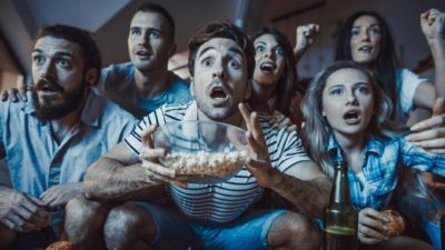 A group of young people sit together watching a television very intently with wide-mouthed, awed expressions while one holds a large bowl of popcorn with a bottle of beer in the foreground.