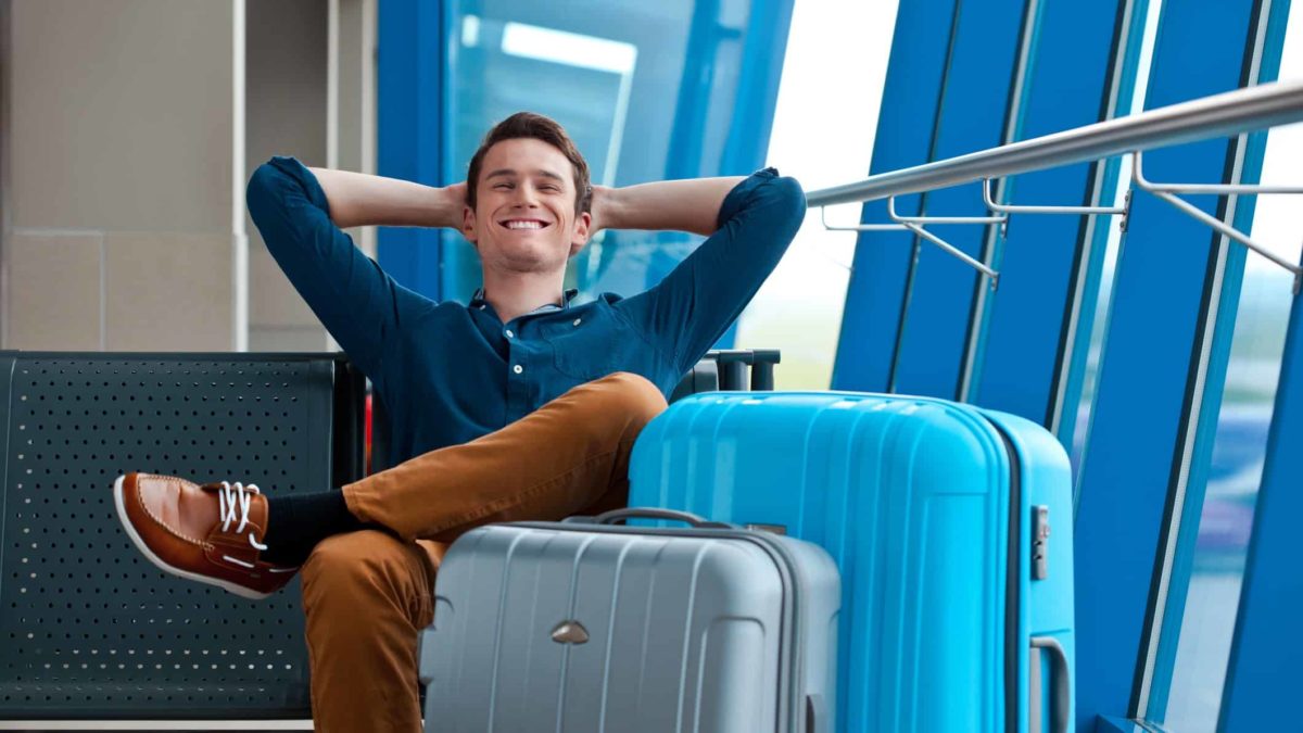 a young man rests back into his hands behind his head with a wide smile and his eyes closed as he sits with two large suitcases in what looks to be an airport or transit destination.