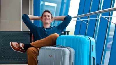 a young man rests back into his hands behind his head with a wide smile and his eyes closed as he sits with two large suitcases in what looks to be an airport or transit destination.