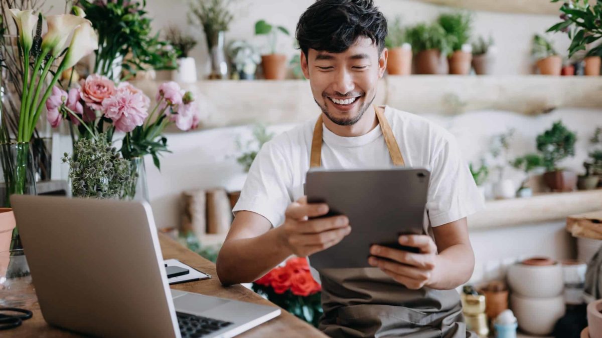 A florist gets some good news on his laptop and tablet, a big smile on his face as he is surrounded by flowers.