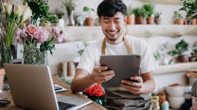 A florist gets some good news on his laptop and tablet, a big smile on his face as he is surrounded by flowers.