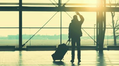 A person holding a suitcase waves goodbye as the sun sets outside the airport terminal.