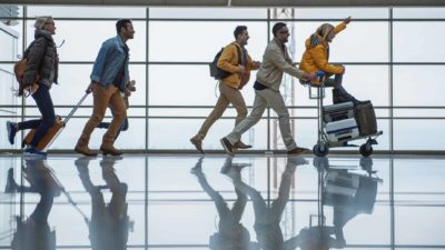 a group of people walk rapidly in a line with airport trolleys and carting baggage as they appear to excitedly set off at the airport on a trip.