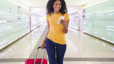 a young woman looks at here phone as she strides out in an airport dragging her wheelie bag behind her and smiling widely.